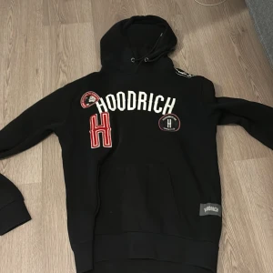 Svart hoodie från Hoodrich - Säljer en svart hoodie från Hoodrich med coola detaljer som broderade loggor och text på framsidan. Den har en klassisk känguruficka och justerbar huva. Perfekt för en avslappnad och stilren look!