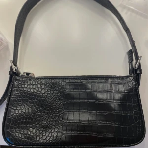 Svart axelväska från Gina Tricot  - En svart väska i fejk krokodil skinn från Gina Tricot. Den har ett justerbart axelband.  Säljer för att den inte längre används! Den är i bra skick💕nypris 300