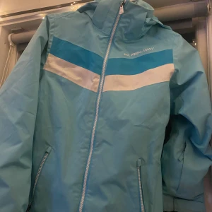 Turkos vindjacka med dragkedja - Säljer en snygg turkos vindjacka med dragkedja och huva. Jackan har en cool design med vita och blå ränder över bröstet. Den har praktiska fickor med knappar och en meshficka på insidan. Perfekt för vår och höst!