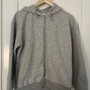Grå hoodie med dragkedja - Säljer en klassisk grå hoodie med dragkedja framtill. Den är supermjuk och perfekt för kyliga dagar. Hoodien har långa ärmar och en bekväm passform. Perfekt för både vardag och mysiga hemmakvällar! Säljes för 100kr. Köptes på Gina, är i bra kvalitet 