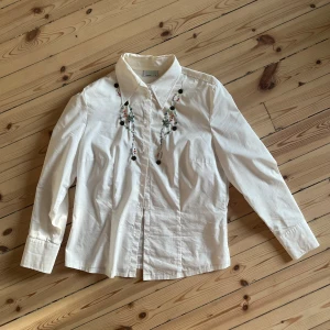 Vit blus med broderade detaljer - Säljer en superfin vit blus med långa ärmar och snygga broderade detaljer i olika färger på framsidan. Perfekt för både vardag och fest! Blusen har en klassisk krage och knappar framtill. Materialet känns som bomull, vilket gör den bekväm att bära.