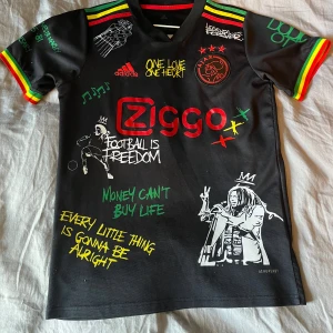 Svart fotbollströja med Bob Marley-tema - Säljer en svart fotbollströja från Adidas med Bob Marley-tema. Tröjan har färgglada detaljer i rött, gult och grönt på ärmarna och runt halsen. Den har flera tryck med Bob Marley-citat och bilder, samt texten 'Football is Freedom' och 'Money Can't Buy Life'. På baksidan finns en stor bild av Bob Marley med texten 'Live the life you love, love the life you live'.