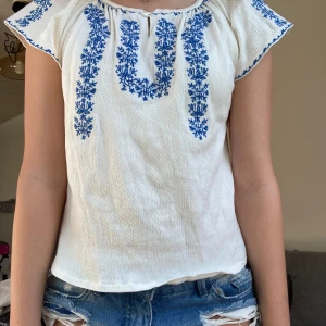 Vit blus med blå broderi - Säljer en superfin vit blus med blått broderi. Blusen har korta ärmar och en liten knappdetalj vid halsen. Perfekt för sommaren och kan stylas både uppklätt och nedklätt. Den är i bra skick och väldigt bekväm att ha på sig.