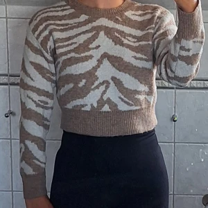 Beige och vit stickad tröja - Säljer en superfin stickad tröja i beige och vitt. Den har ett coolt zebramönster och är croppad, vilket gör den perfekt för både höst och vinter. Tröjan har långa ärmar och är riktigt mysig att ha på sig. Perfekt för att hålla sig varm och snygg under de kallare månaderna!
