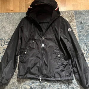 Svart vindjacka från Moncler - Säljer en snygg svart vindjacka från Moncler. Jackan har en dragkedja framtill, två fickor med knappar och en justerbar huva med snören. Den är perfekt för vår och höst och har en normal passform. Moncler-loggan sitter på vänster ärm.
