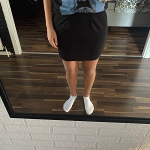 Svart kjol med knytband - Säljer en snygg svart kjol som är perfekt för både vardag och fest. Kjolen har en enkel och stilren design med knytband i midjan för justerbar passform. Den är kort och passar bra till olika toppar och skor. Perfekt för alla säsonger!