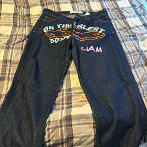 Mörkblå jeans med broderad text - Säljer ett par mörkblå jeans från Catch i storlek L. De har en cool broderad text och design på framsidan med orden 'ON THE ALERT' och 'LIAM' i olika färger. Jeansen har en klassisk femficksdesign och är i bra skick. Perfekta för en avslappnad och trendig look!