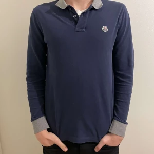 Mörkblå pikétröja från Moncler - Säljer en stilren mörkblå pikétröja från Moncler med grå krage och muddar. Tröjan har långa ärmar och en liten logotyp på bröstet. Perfekt för en avslappnad men ändå snygg look. Storlek m slim fit.