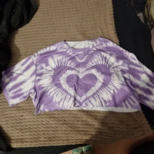 Lila tie-dye croppad t-shirt - Säljer en cool lila och vit tie-dye croppad t-shirt med ett hjärtmönster i mitten. Perfekt för en avslappnad stil med korta ärmar och en lös passform. Passar bra till jeans eller shorts för en somrig look. Osäker på exakt storlek men skulle gissa på M.
