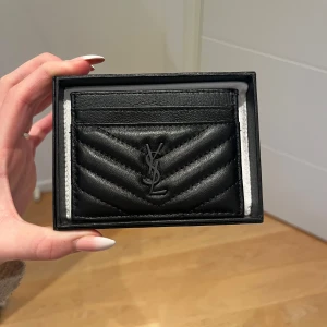 Svart quiltad kortfodral från YSL - Säljer ett stilrent svart kortfodral från YSL i quiltat skinn. Perfekt för att hålla dina kort organiserade med stil. Diskret och elegant design med YSL-logga på framsidan.