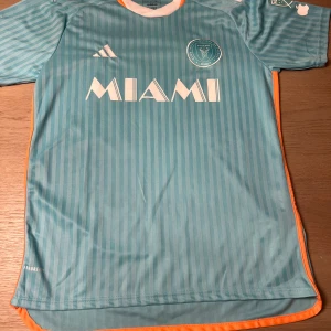 Turkos fotbollströja från Adidas - Säljer en snygg turkos fotbollströja från Adidas med vit text och orange detaljer. Tröjan har korta ärmar och ett randigt mönster. Perfekt för fotbollsfans!