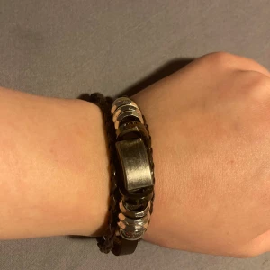 Brunt armband med metalldetaljer  - Snyggt armband i brunt läder med en robust metallkedja i mitten. Perfekt för att ge en stilren touch till din outfit. Armbandet har en flätad design som ger det en unik look.