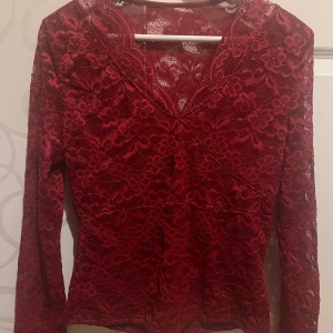 Röd spetsblus - Säljer en elegant röd spetsblus med v-ringning och långa ärmar. Blusen har ett vackert blommigt mönster och är perfekt för en stilfull look. Den är tillverkad i ett mjukt och bekvämt material. Aldrig använd, sitter tajt och man ser inte igenom!