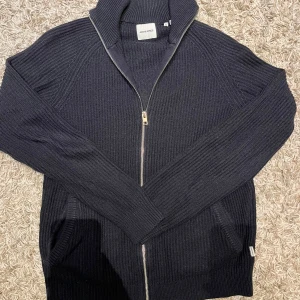 Marinblå stickad kofta från Jack & Jones - Säljer en stilren marinblå stickad kofta från Jack & Jones. Koftan har en praktisk dragkedja framtill och långa ärmar. Perfekt för kyligare dagar. Passar bra till både jeans och chinos.