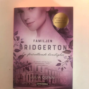 Familjen Bridgerton: En förtrollande hemlighet - Boken 'Familjen Bridgerton: En förtrollande hemlighet' av Julia Quinn är en del av den populära Bridgerton-serien. Den har ett vackert omslag i lila toner med en bild av en kvinna och ett hus i bakgrunden. Utvald av Simona Ahrnstedt, boken lovar en förtrollande berättelse i en historisk miljö.