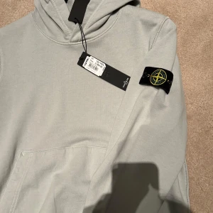 Grå hoodie från Stone Island - Säljer en stilren hoodie från Stone Island med deras ikoniska märke på ärmen. Den är helt ny, Den är i storlek 152 och är 100% äkta igår NK. 