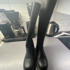 Svarta stövlar med dragkedja  - Snygga svarta stövlar i skinn med grov sula och dragkedja på sidan. De har en dekorativt spenne och är fodrade med päls för extra värme. Perfekta för kyligare dagar. Använd få tal gånger