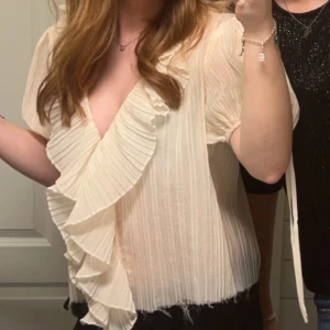 Vit blus med volangdetaljer - Säljer en elegant vit blus med v-ringning och volangdetaljer framtill. Blusen har korta ärmar och är perfekt för en stilren look. Passar bra till både jeans och kjol. Från Zara 