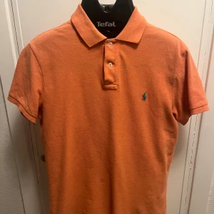 Orange pikétröja från Ralph Lauren - Säljer en snygg orange/persiko färgad pike tröja från Ralph Lauren. Tröjan har en turkos logga och är slim fit i storleken M. Pris kan diskuteras
