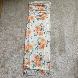 Blommig axelbandslös långklänning M-L stretch oanvänd  - Snygg axelbandslös långklänning med ett vackert blommigt mönster i orange och grönt på en vit bakgrund. Perfekt för sommarens festligheter!