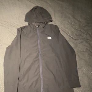 North Face Tunn Jacka - Säljer en stilren svart vindjacka från The North Face. Jackan har en praktisk huva och dragkedja framtill. Perfekt för blåsiga dagar och har en diskret logga på bröstet. Passar bra för utomhusaktiviteter och vardagsbruk.