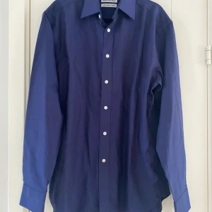 Mörkblå skjorta från The Shirt Factory - Snygg mörkblå skjorta från The Shirt Factory, designad av Linnéa Braun. Tillverkad i 100% bomull med lättstruken finish. Skjortan har långa ärmar och knappar framtill. Perfekt för en stilren look. Ny marinblå skjorta i strl 37 från The Shirt Factory.  Ny, aldrig använd.  Mått Över bröstet: ca 54 cm Längd från axeln ner: ca 75 cm  100% bomull   Kommer från ett djur och rökfritt hem. 