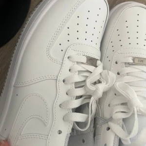 Vita Nike Air Force sneakers - Klassiska vita Nike Air Force sneakers med snörning och perforerade detaljer för ventilation. Dessa sneakers har en ikonisk design med en robust sula och Nike-loggan på sidan. Perfekta för en stilren och sportig look.