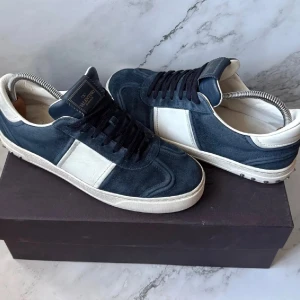 Blå och vita sneakers från Valentino - Snygga sneakers från Valentino i blå mocka med vita detaljer. Skorna har en klassisk design med snörning och en vit sula. Perfekta för en stilren look.