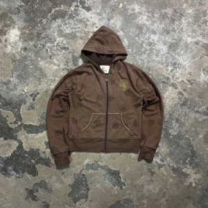 zip up hoodie - Säljer en y2k brun zip up hoodie med dragkedja och mönster av logotyper. Tröjan har en ficka framtill och en broderad detalj på bröstet. as cool🔥 