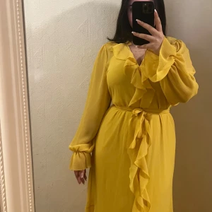 Gul klänning med volangdetaljer - Säljer en solgul klänning med vackra volangdetaljer och knytband i midjan. Klänningen har långa ärmar och en omlott-design framtill som ger en elegant look. Perfekt för att lysa upp garderoben!