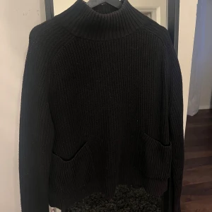 Svart stickad tröja  - Säljer en svart stickad tröja från Monki med hög krage och långa ärmar. Tröjan har en ribbad design och praktiska fickor framtill. Även en slit i ryggen. Perfekt för kyliga dagar. Hör av dig vid intresse!