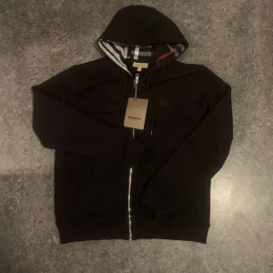 Svart huvtröja från Burberry - Säljer en stilren svart huvtröja från Burberry med dragkedja och klassiskt rutigt mönster i fodret. Tröjan har långa ärmar och en bekväm passform. Perfekt för en avslappnad look.