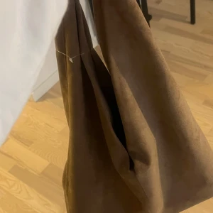 Brun slutsåld axelväska i mocka från ginatricot - Snygg och stilren brun axelväska i mocka. Perfekt för att bära med sig det nödvändigaste med stil. Väskan har en enkel design och är i one size. Aldrig använd, slutsåld. 