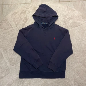 Mörkblå hoodie från Polo Ralph Lauren - Säljer en snygg mörkblå hoodie från Polo Ralph Lauren med det klassiska röda logotypbroderiet på bröstet. Tröjan har en bekväm huva och långa ärmar, perfekt för kyliga dagar. Passar både till vardags och avslappnade tillfällen. Passar 14-16