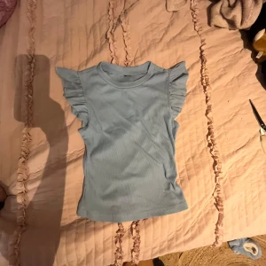 Ljusblå ribbad topp med volangärmar - Säljer en söt ljusblå ribbad topp med volangdetaljer på ärmarna. Perfekt för en avslappnad och stilren look. Toppen är ärmlös och har en tight passform.