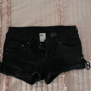 low waist svarta shorts med snörning från H&M - Snygga svarta shorts från H&M i denim med cool snörning på sidorna. Perfekta för en trendig look. De har en klassisk femficksdesign och knappgylf.