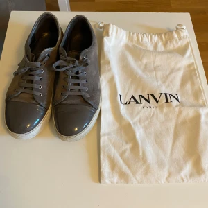 Grå sneakers från Lanvin - Snygga grå sneakers från Lanvin med snörning och en stilren design. Skorna har en glansig tådel och kommer med en praktisk tygpåse för förvaring. Skorna är i den eftertraktade färgen elpiano grey. Hör av er vid funderingar!😃