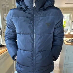 Mörkblå Dunjacka - Säljer en snygg mörkblå dunjacka med dragkedja och huva från Guess. Fint skick men har ett litet hål i fickan. 