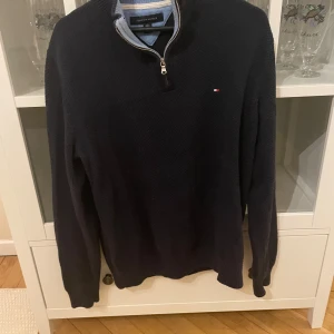 Mörkblå tröja med dragkedja från Tommy Hilfiger - Säljer en stilren mörkblå tröja från Tommy Hilfiger med dragkedja vid halsen. Tröjan har långa ärmar och en liten logotyp på bröstet. Perfekt för en avslappnad och snygg look.
