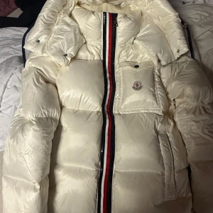 Vit dunjacka från Moncler - Säljer en snygg vit dunjacka från Moncler med en iögonfallande randig dragkedja i rött, vitt och blått. Jackan har en praktisk ficka med Moncler-logga på bröstet och en hög krage för extra värme. Perfekt för kyliga dagar!