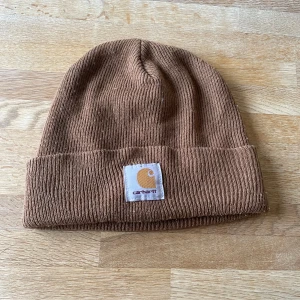 Brun ribbad mössa från Carhartt - Säljer en snygg brun ribbad mössa från Carhartt. Den har en uppvikt kant och en broderad logotyp framtill. Perfekt för kyliga dagar.