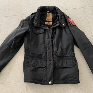 Svart jacka från Parajumpers - Säljer en svart jacka från Parajumpers med coola detaljer som en broderad patch på ärmen och flera fickor framtill. Jackan har en dragkedja och knappar för stängning samt en hög krage. Perfekt för höst och vår!