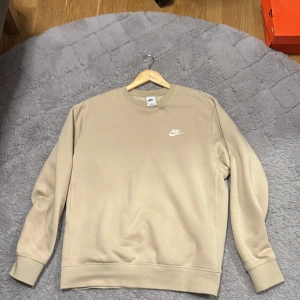Beige sweatshirt från Nike - Säljer en stilren beige sweatshirt från Nike med broderad logga på bröstet. Tröjan har långa ärmar och ribbade muddar vid ärmslut och nederkant. Perfekt för en avslappnad look.