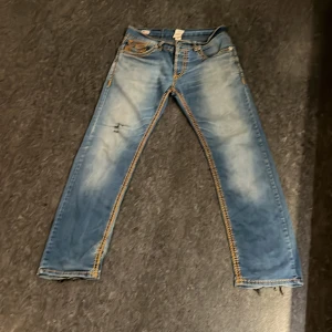Blå jeans från True Religion - Säljer ett par blå jeans från True Religion med slitna detaljer och kontrastsömmar. Jeansen har en klassisk femficksdesign och broderade bakfickor. Perfekta för en avslappnad stil. Dom har lagade hål vid skrevet och på knät! Fråga gärna om bilder för mer!
