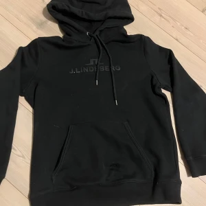Svart hoodie från J.Lindeberg - Säljer en stilren svart hoodie från J.Lindeberg. Den har en klassisk design med dragsko i huvan och en praktisk magficka. Perfekt för en avslappnad look. den är knappt använd och i storleken L men skulle säga att den mer passar M
