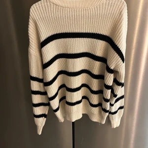 Randig stickad tröja - Säljer en mysig stickad tröja i beige med svarta ränder. Tröjan har en avslappnad passform och långa ärmar, perfekt för kyligare dagar. Den ribbade strukturen ger en extra touch av stil.
