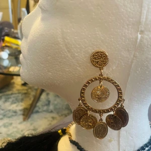 Guldörhängen med hängande mynt - Stora och iögonfallande guldörhängen med flera hängande mynt. Designen har en cirkulär bas med detaljerade mönster och flera små mynt som dinglar nedanför. Perfekt för att ge en bohemisk touch till din stil.