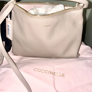   axelväska från Coccinelle i rosette färg - Snygg rosette  axelväska från Coccinelle i skinn. Väskan har en elegant design med en praktisk dragkedja upptill. Perfekt för att bära dina dagliga nödvändigheter med stil. Den kommer med en justerbar axelrem för bekväm användning.