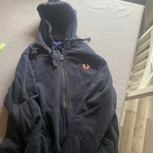 Svart hoodie från Fred Perry - Säljer en svart hoodie från Fred Perry med dragkedja och huva. Den har en broderad logga på bröstet och snörning vid huvan. Perfekt för en avslappnad stil.