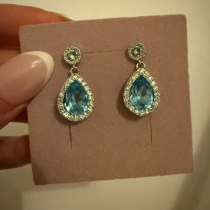 Blå droppformade örhängen med stenar - Eleganta örhängen med en stor blå droppformad sten omgiven av gnistrande stenar. Fästet är runt med en mindre sten. Perfekta för att ge en touch av glamour.